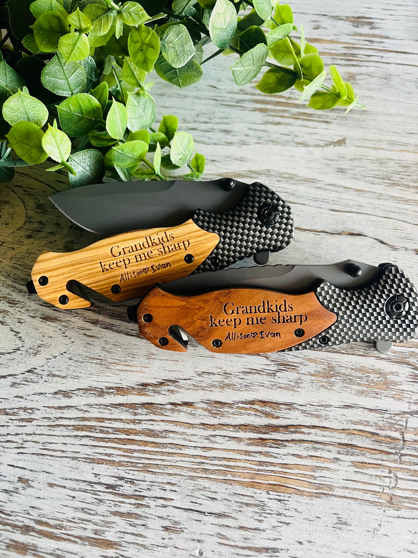 Two folding pocket knives with wooden handles, one with the blade open displaying the engraved text on the handle, the other with the blade closed. Set against a wooden background with green leaf accents.