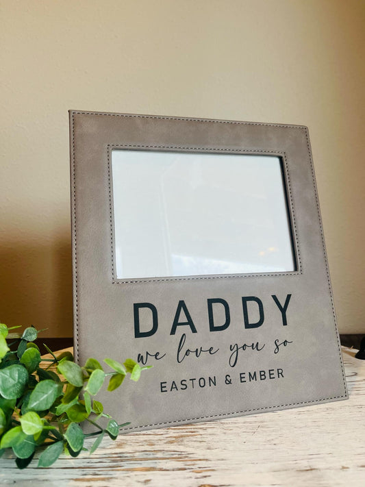 A gray leather picture frame with the text 'DADDY we love you so EASTON & EMBER' engraved on the bottom. The frame is displayed on a wooden surface with a plant to the side.