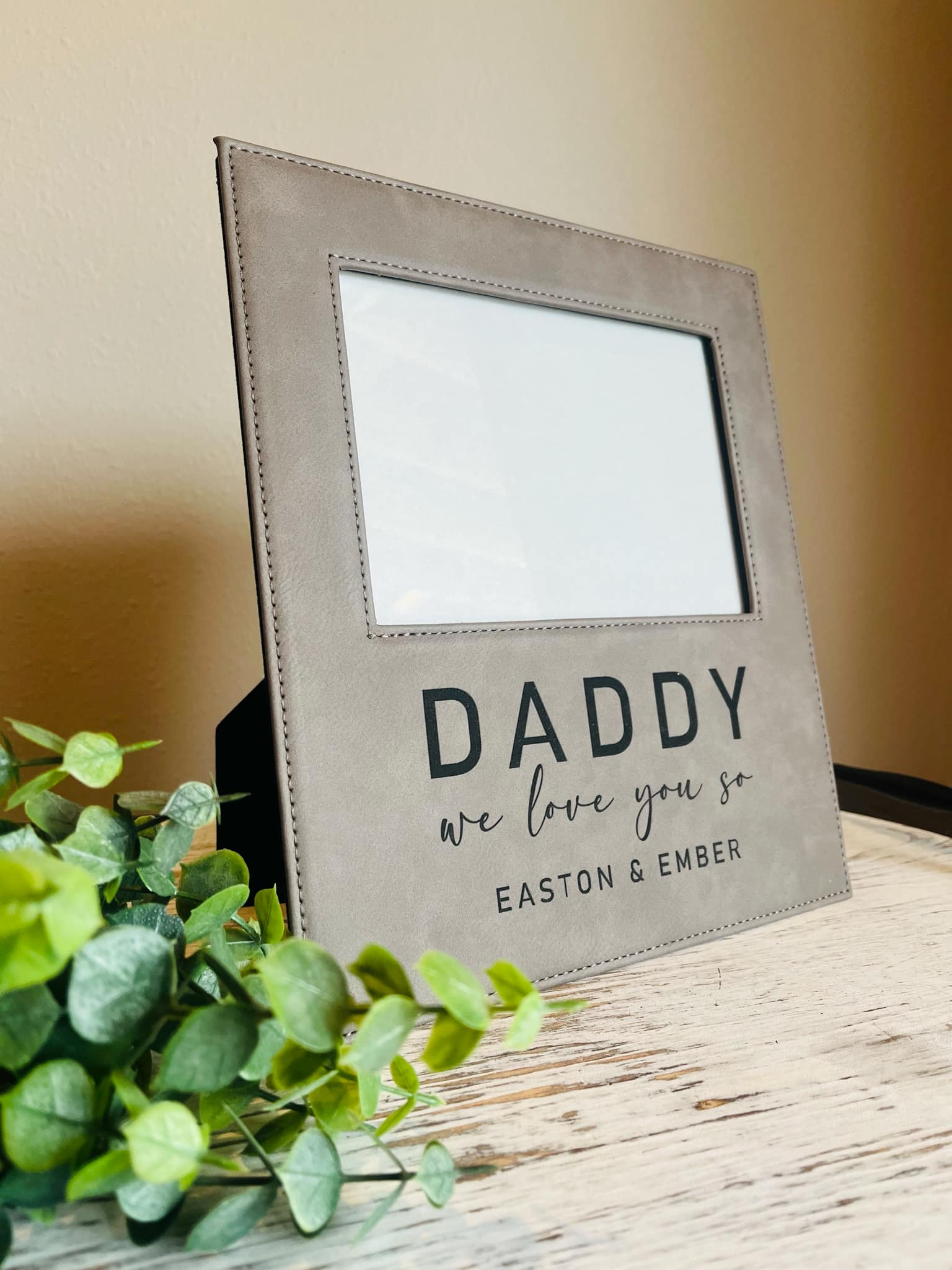 A gray leather picture frame with the text 'DADDY we love you so EASTON & EMBER' engraved on the bottom. The frame is displayed on a wooden surface with a plant to the side.