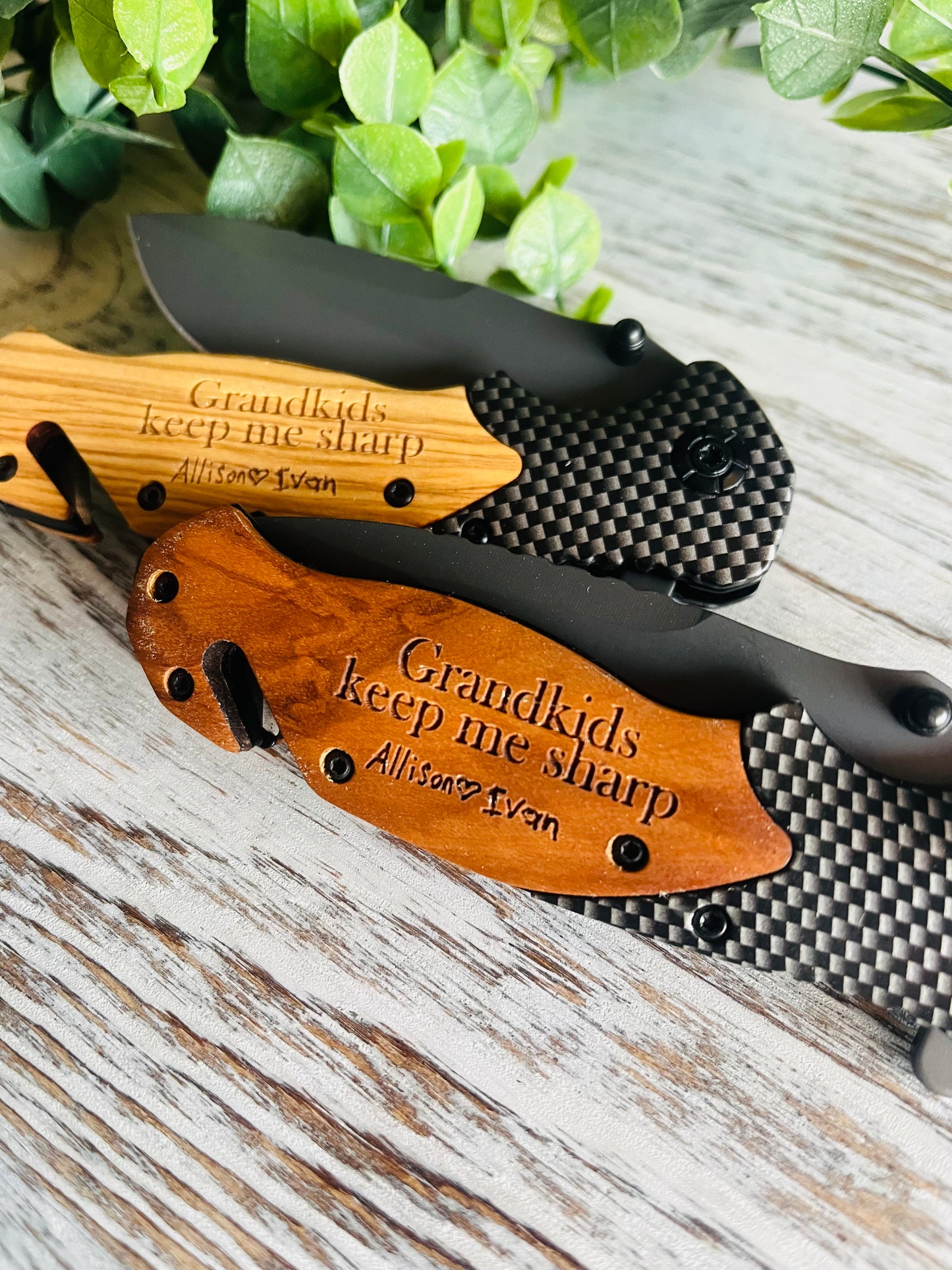 Two folding pocket knives with wooden handles, one with the blade open displaying the engraved text on the handle, the other with the blade closed. Set against a wooden background with green leaf accents.