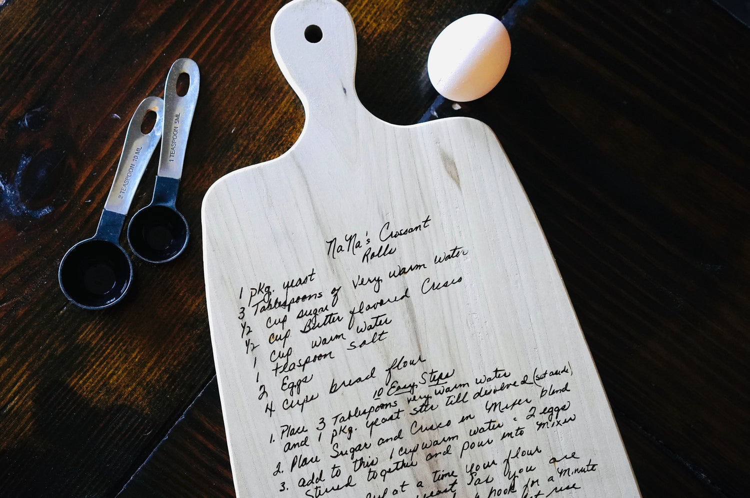A wooden cutting board with a personalized engraved recipe, displayed on a table with a pair of scissors and a magnifying glass in the background.
